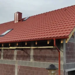 Dom w trakcie budowy z czerwoną dachówką, widoczne okna dachowe, komin i orynnowanie w kolorze dachu, ściany z pustaków ceramicznych z elementami żelbetowymi.