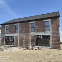 Surowy stan dwukondygnacyjnego domu z ciemnej cegły z zamontowanymi oknami, widoczne materiały budowlane na placu budowy i sąsiedni dom w tle.