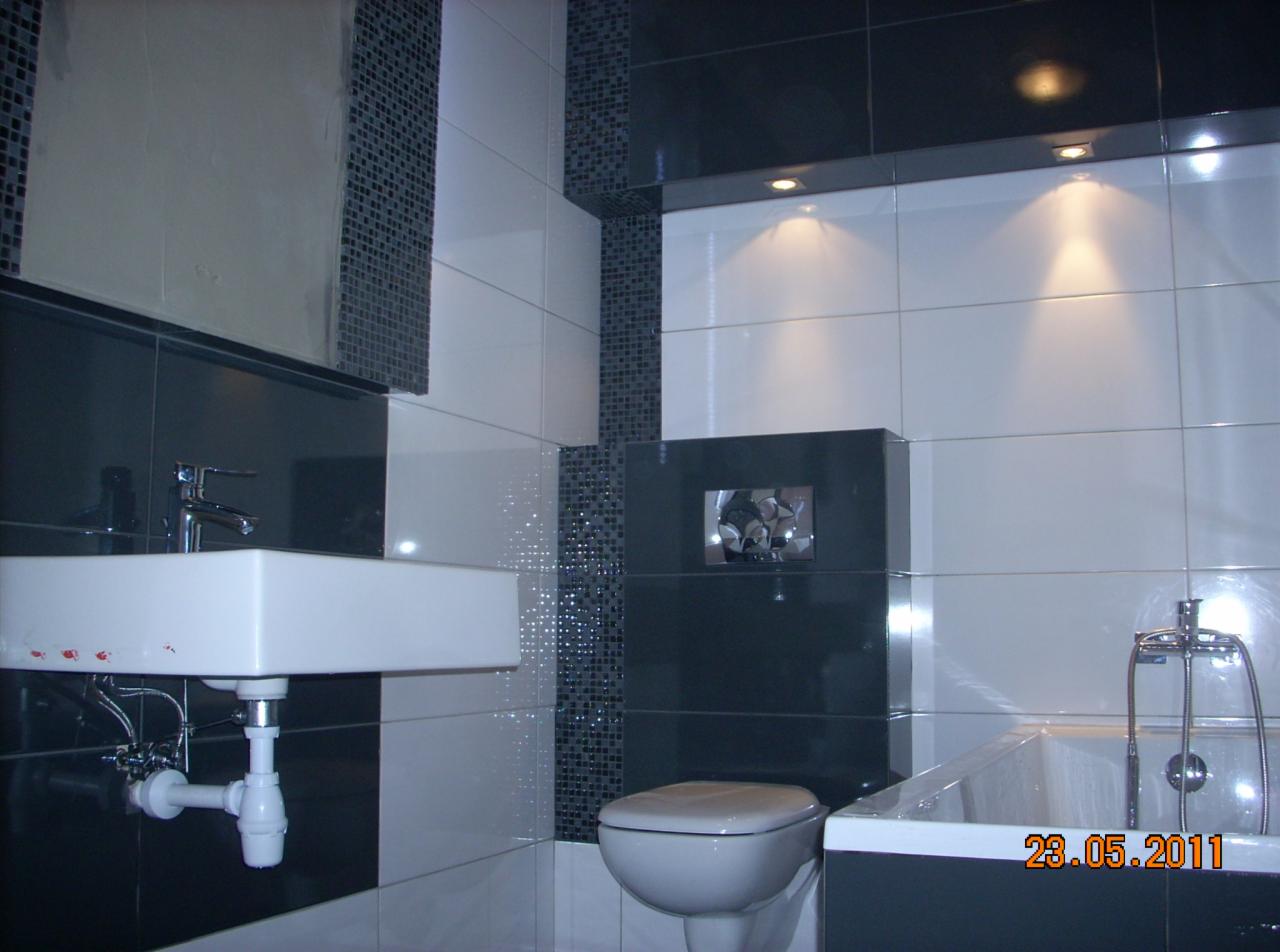 Nowoczesna łazienka w bieli i czerni z mozaiką, umywalką na ścianie i podświetlanym sufitem. Widoczna wanna i toaleta. Ukończone wykończenie łazienki.