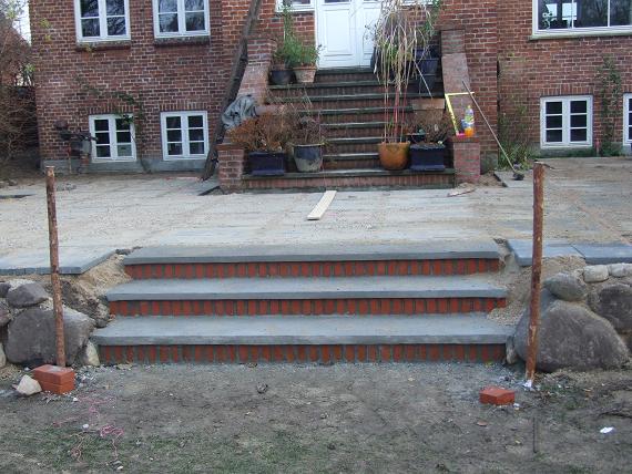 Budowa schodów wejściowych z cegły klinkierowej i betonu prowadzących do domu z czerwonej cegły. Widoczne fragmenty podjazdu z piasku i kostki oraz elementy konstrukcyjne.