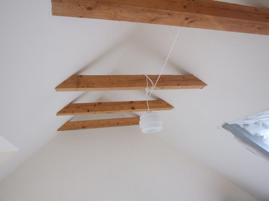 Drewniane belki stropowe w kształcie trójkątów podtrzymujące białą lampę wiszącą na białym sznurze, widok z dołu.