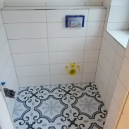 Wnętrze łazienki w trakcie remontu, widoczne białe płytki ścienne z metalową listwą, podtynkowy stelaż WC z niebieską ramką, żółte rury instalacyjne oraz podłoga wyłożona wzorzystymi płytkami...