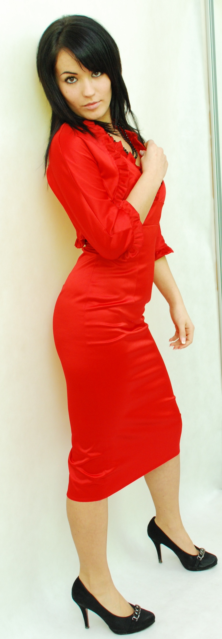 Kobieta w czerwonej, dopasowanej sukience i czarnych szpilkach z ozdobnym łańcuszkiem, pozująca na jasnym tle.