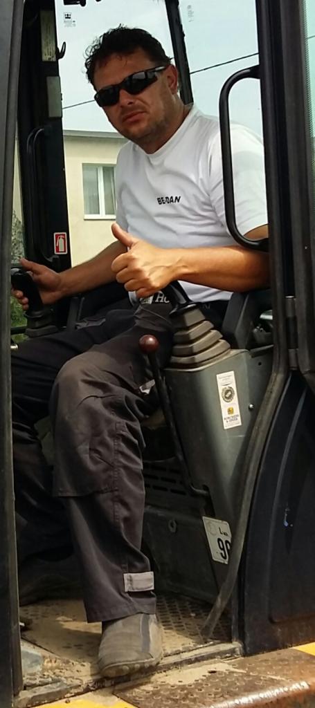 Operator w kabinie koparki, ubrany w białą koszulkę z napisem, kciuk w górę. Widoczne elementy sterowania i szyba kabiny. Operator w okularach przeciwsłonecznych.