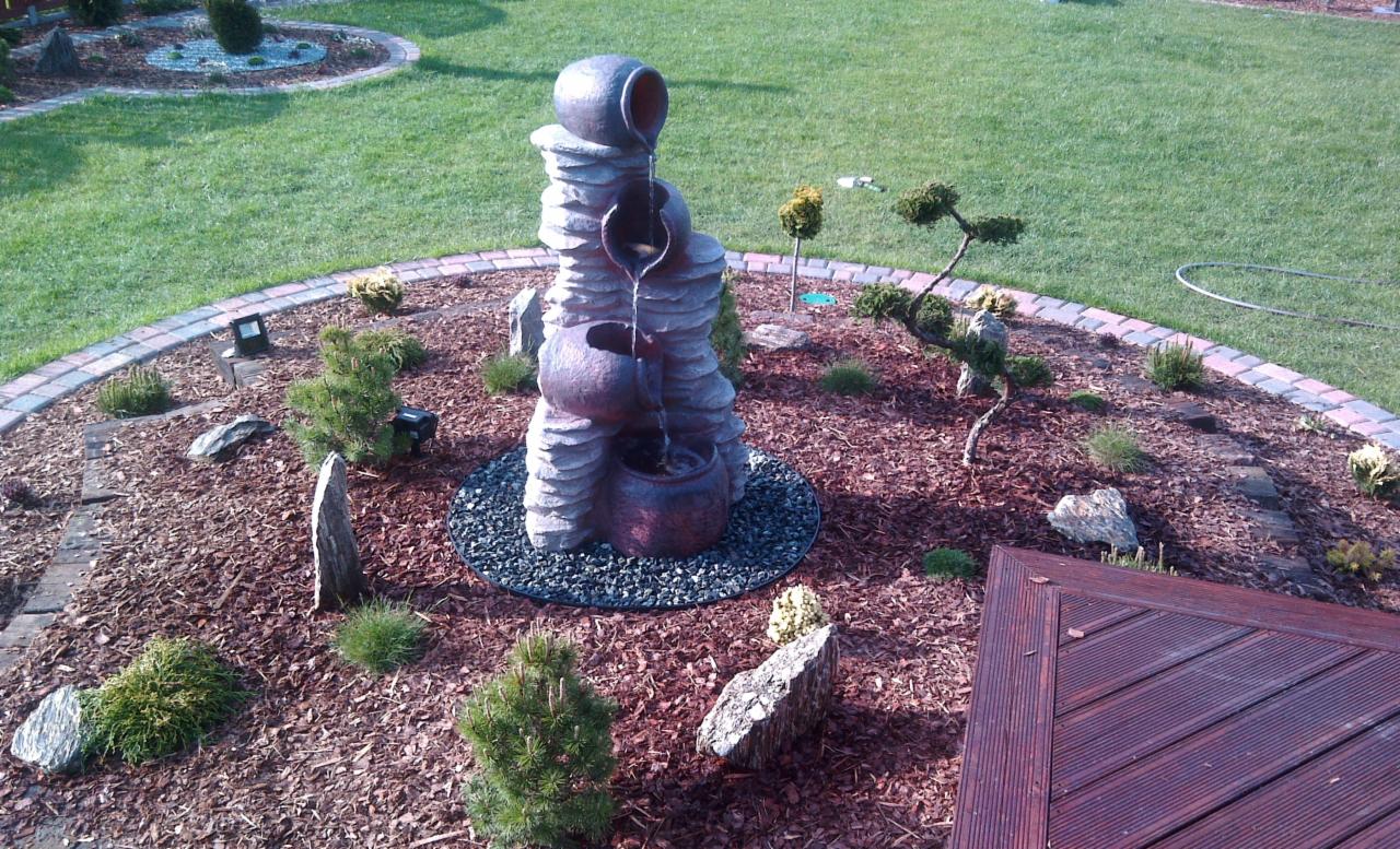 Ogród z dekoracyjną, trójpoziomową fontanną w kształcie dzbanów, otoczoną korą i kamieniami, z widocznym fragmentem drewnianego tarasu w rogu