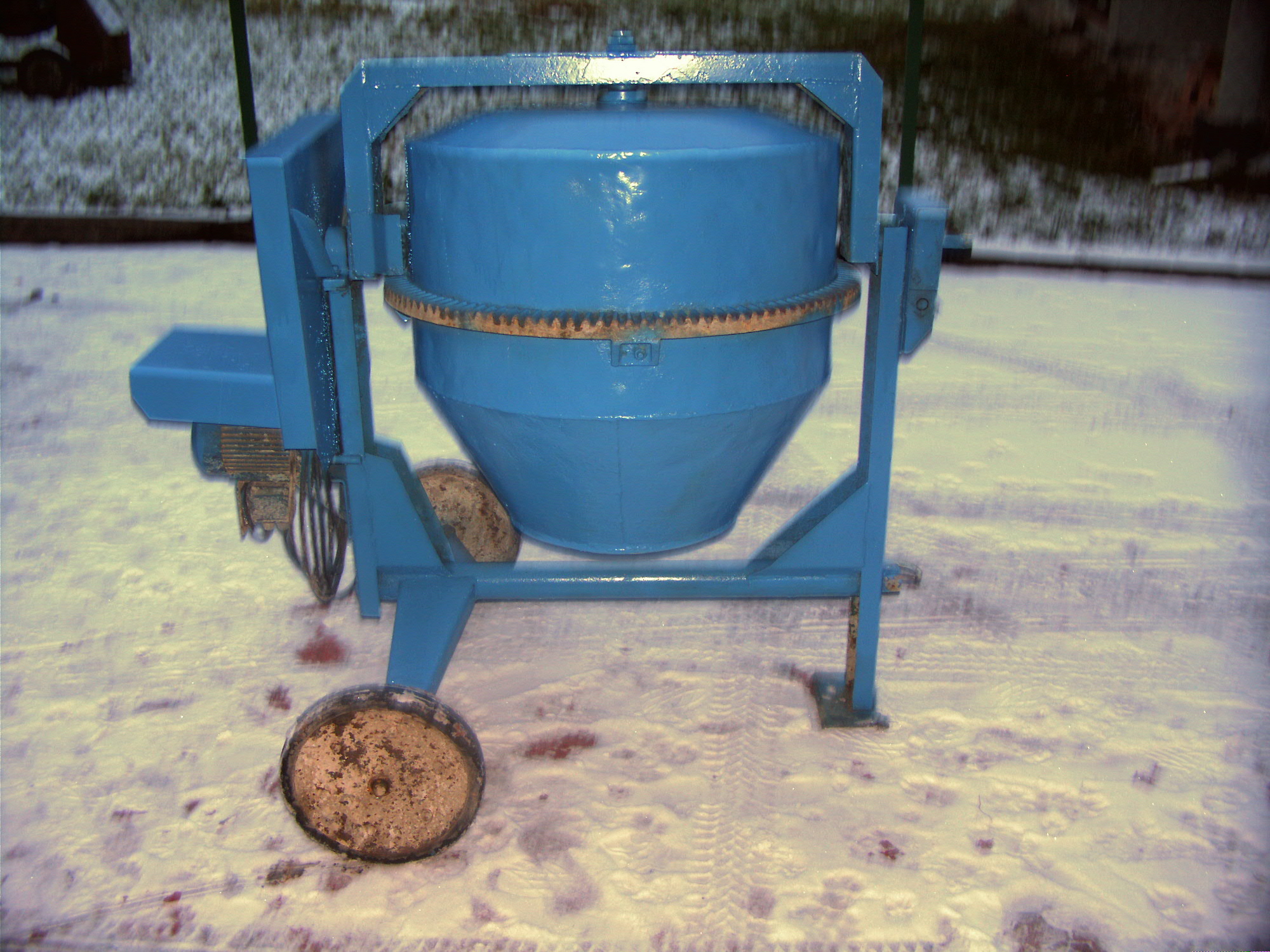 Niebieska betoniarka stojąca na śniegu, widoczne ślady opon i elementy konstrukcyjne urządzenia.