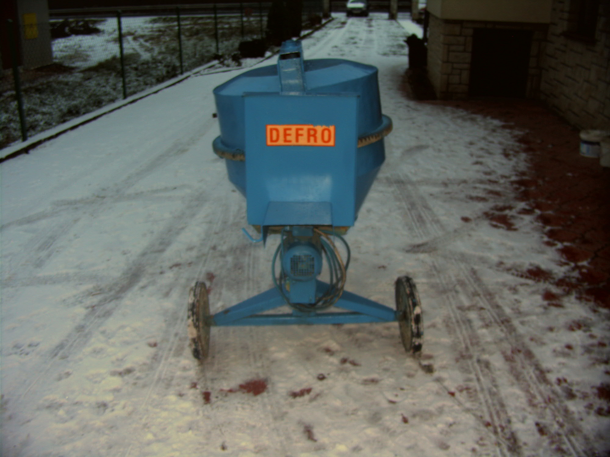 Niebieska betoniarka marki DEFRO na kołach, stojąca na zaśnieżonym podłożu przed budynkiem w zimowy dzień.