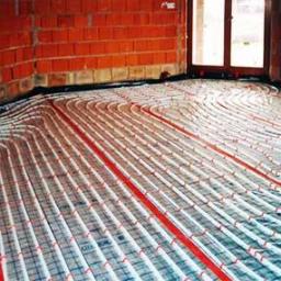 Instal-spaw usługi hydrauliczne i spawalnicze Sebastian Lewczuk - Ułożone rury ogrzewania podłogowego na folii izolacyjnej, widoczne ceglane ściany w surowym stanie i fragment okna. Etap instalacji przed wylewką.