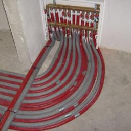 Instal-spaw usługi hydrauliczne i spawalnicze Sebastian Lewczuk - Rozdzielacz ogrzewania podłogowego z licznymi szarymi i czerwonymi rurami ułożonymi w podłodze, wnęka w ścianie, surowy stan wykończenia.
