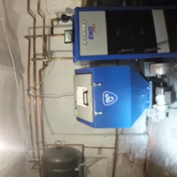 Zamontowany piec Eko Plus z rurami miedzianymi i zbiornikiem w kotłowni. Widoczne elementy instalacji grzewczej. Białe ściany, surowy stan.