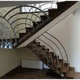 Schody z metalową balustradą o łukowym wzorze, drewniane stopnie, białe ściany. Widok z dołu na eleganckie wykończenie wnętrza domu.