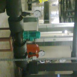 Instal-spaw usługi hydrauliczne i spawalnicze Sebastian Lewczuk - Instalacja przemysłowa z rurami, manometrami i elementami w kolorach: czarnym, zielonym i czerwonym. Pracownik w żółtej kamizelce w tle.