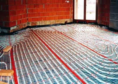 Ułożone rury ogrzewania podłogowego na folii izolacyjnej, widoczne ceglane ściany w surowym stanie i fragment okna. Etap instalacji przed wylewką.