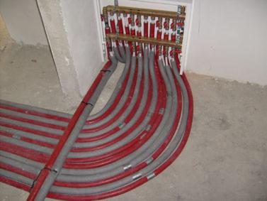 Rozdzielacz ogrzewania podłogowego z licznymi szarymi i czerwonymi rurami ułożonymi w podłodze, wnęka w ścianie, surowy stan wykończenia.