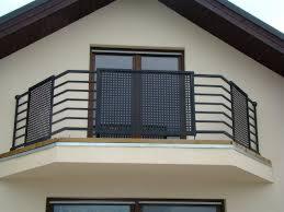 Czarna, metalowa balustrada balkonowa z dekoracyjnym wypełnieniem na balkonie budynku z beżową elewacją i białymi oknami. Ujęcie z dołu, centralne.