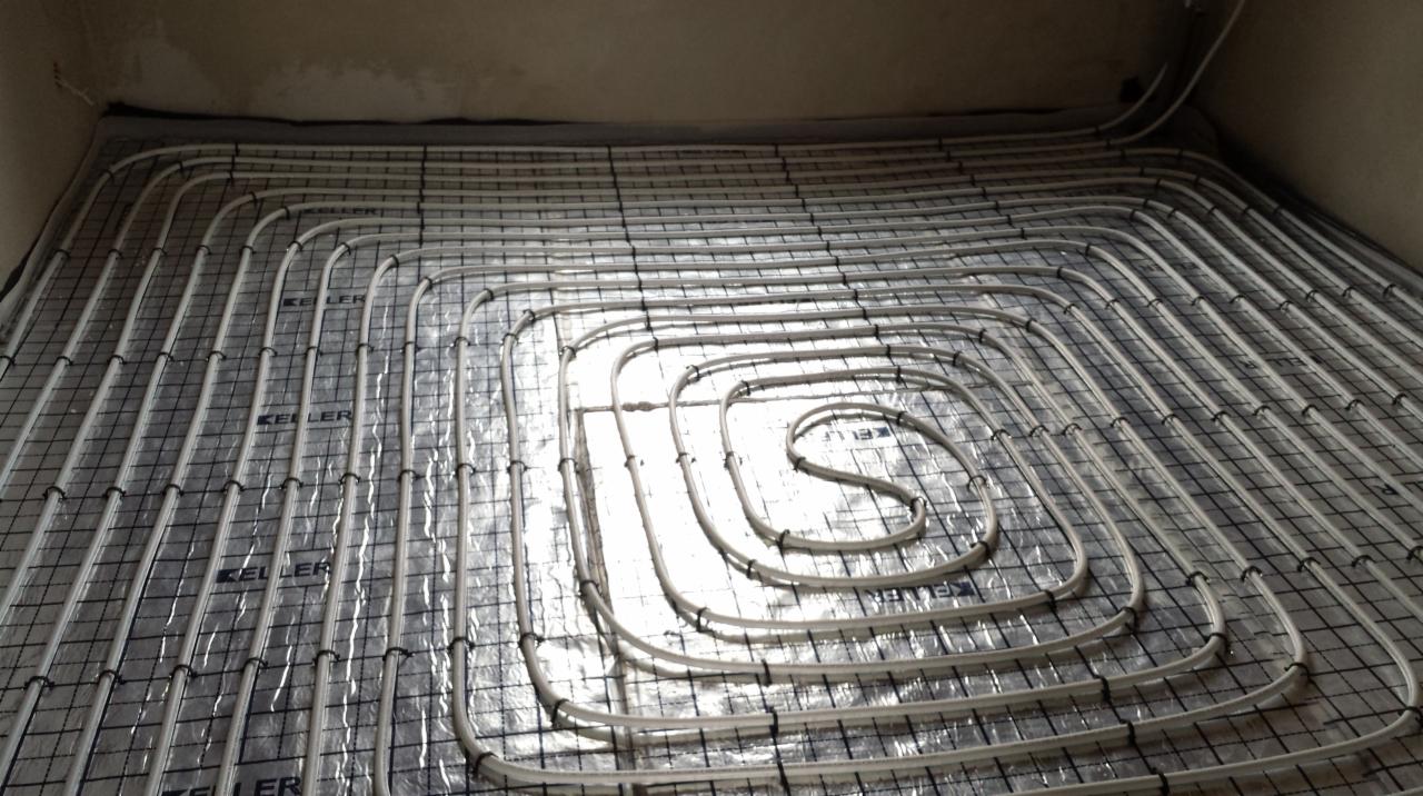 Białe rury ogrzewania podłogowego ułożone spiralnie na srebrnej folii z czarną siatką, przymocowane czarnymi opaskami, gotowe do zalania wylewką.