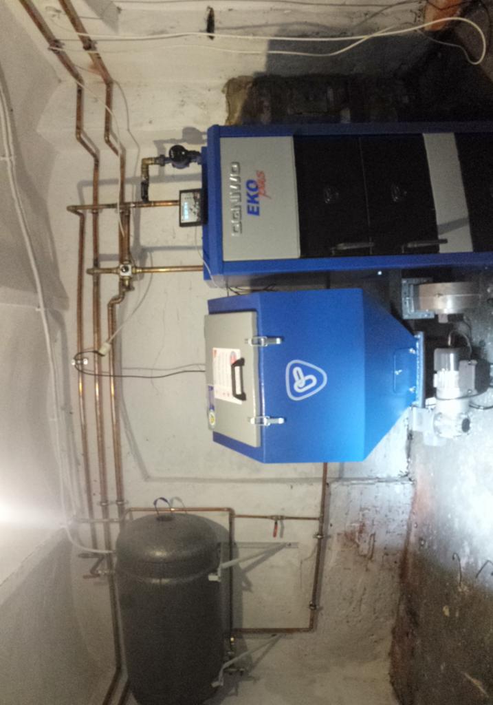 Zamontowany piec Eko Plus z rurami miedzianymi i zbiornikiem w kotłowni. Widoczne elementy instalacji grzewczej. Białe ściany, surowy stan.
