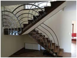 Schody z metalową balustradą o łukowym wzorze, drewniane stopnie, białe ściany. Widok z dołu na eleganckie wykończenie wnętrza domu.