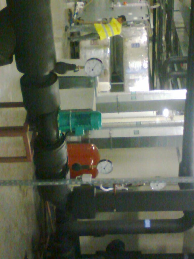 Instalacja przemysłowa z rurami, manometrami i elementami w kolorach: czarnym, zielonym i czerwonym. Pracownik w żółtej kamizelce w tle.