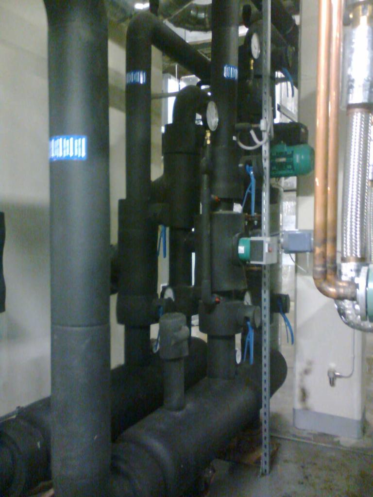 Czarne, zaizolowane rury instalacji przemysłowej z manometrami, zaworami i silnikiem elektrycznym, w hali z metalową konstrukcją pod sufitem i miedzianymi rurami.