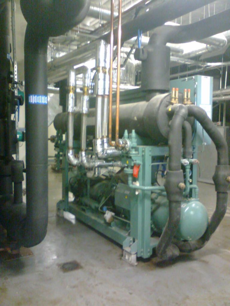 Kompleksowa instalacja przemysłowa chłodnicza: rury, zawory, agregat w hali. Widoczne rury miedziane i czarne izolowane. Elementy pomalowane na zielono.