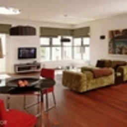 Jasne wnętrze salonu z podłogą z paneli w ciepłym odcieniu brązu, czerwone krzesła wokół okrągłego stolika kawowego z czarnym blatem, zielona sofa z dekoracyjną narzutą, telewizor na niskiej szafce...