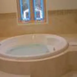 Biała, owalna wanna z hydromasażem, wypełniona wodą, otoczona beżową glazurą i terakotą, z widocznym oknem w tle. Widoczny chromowany kran. Elementy wykończeniowe z gresu.