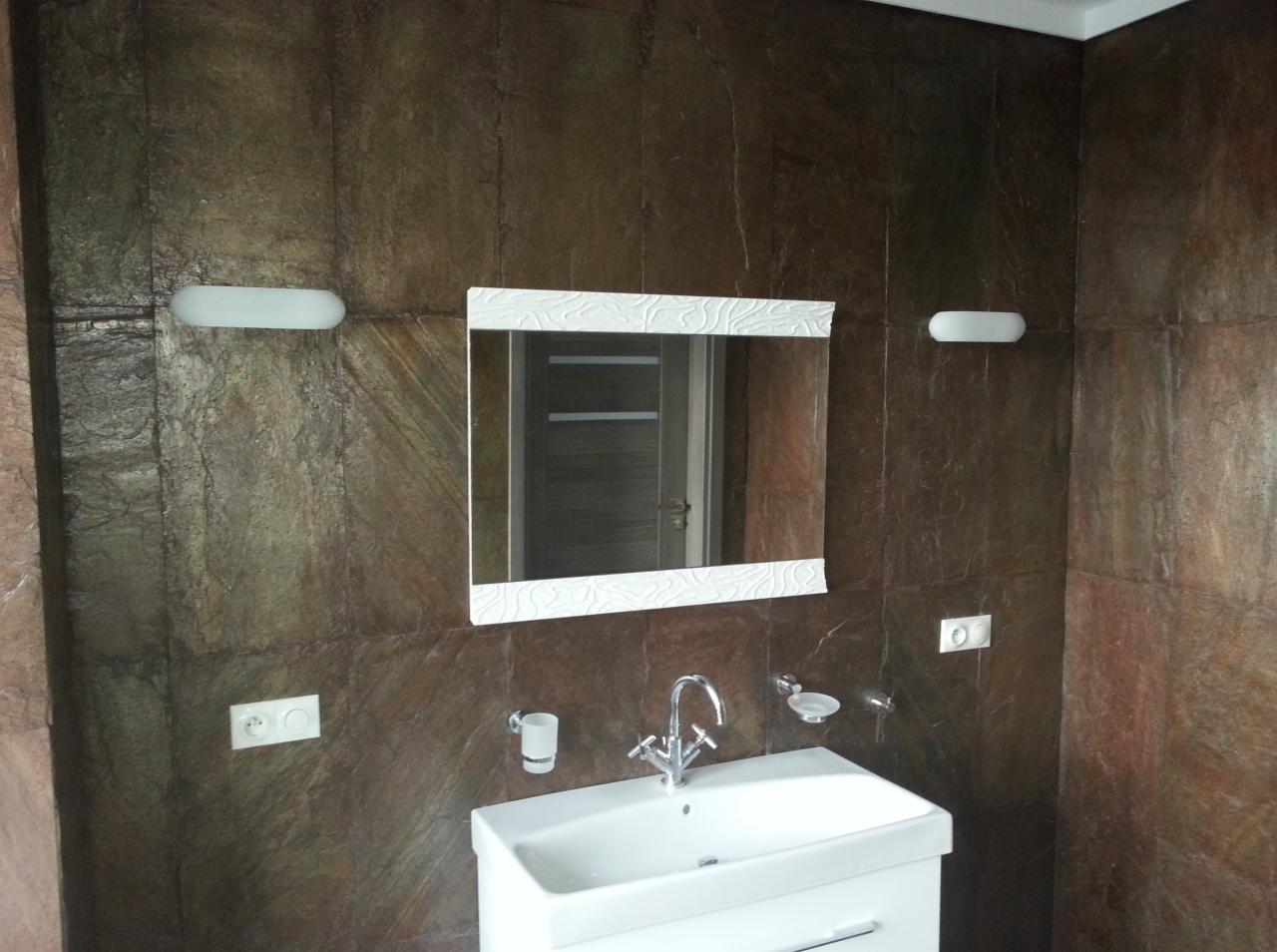 Umywalka z szafką, lustro w białej ramie z wzorem i brązowe płytki imitujące kamień w wykończonej łazience.