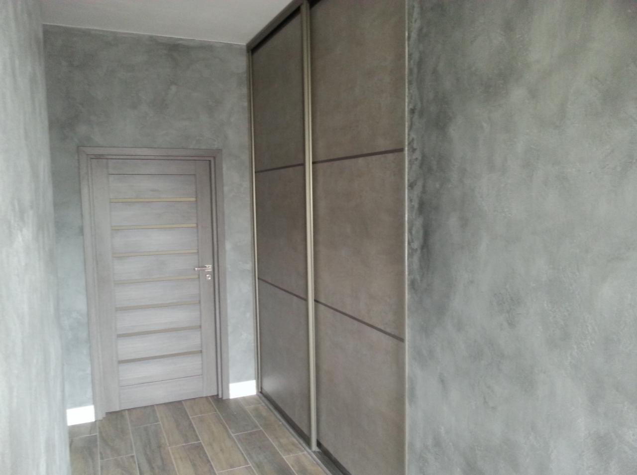 Szara ściana z efektem betonu architektonicznego, drzwi wewnętrzne z poziomymi panelami i przesuwna szafa wnękowa z ciemnymi akcentami, całość w chłodnej tonacji szarości.