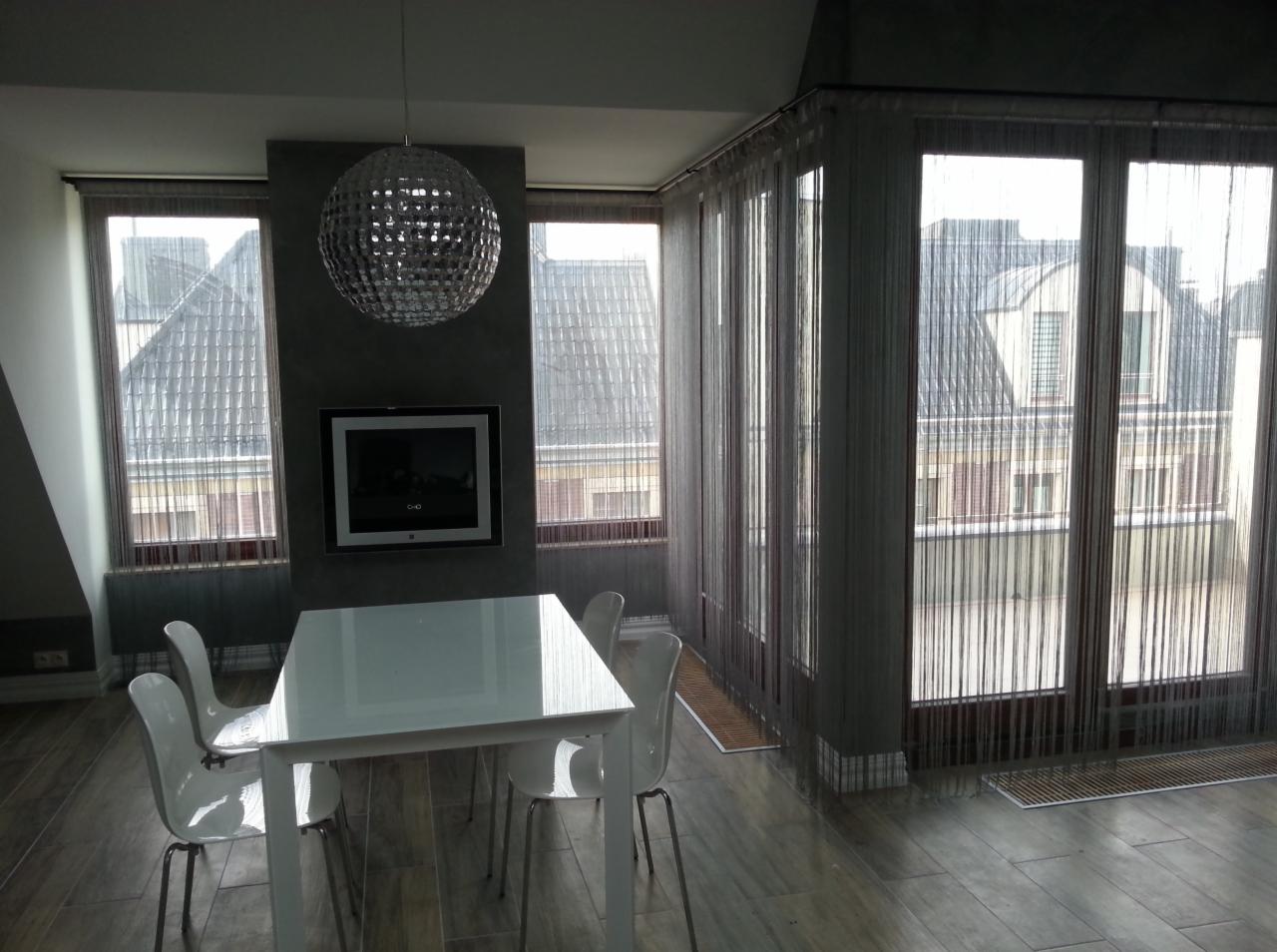 Jasne wnętrze z białym stołem i krzesłami, widok na dachy kamienic przez okna z pionowymi żaluzjami sznurkowymi, srebrna lampa kula nad stołem, telewizor na ścianie.
