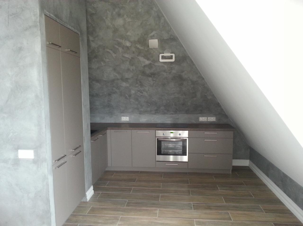 Kuchnia w szarościach z drewnopodobną podłogą, ściany wykończone dekoracyjnym tynkiem imitującym beton, zabudowa kuchenna pod skosem dachu.