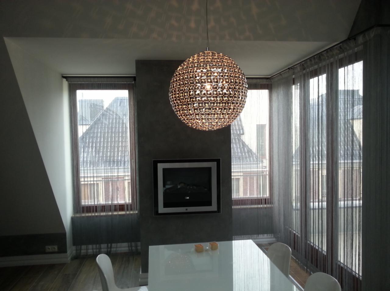 Nowoczesne wnętrze jadalni z kulistą lampą wiszącą, telewizorem wbudowanym w ścianę i dużymi oknami z transparentnymi zasłonami.