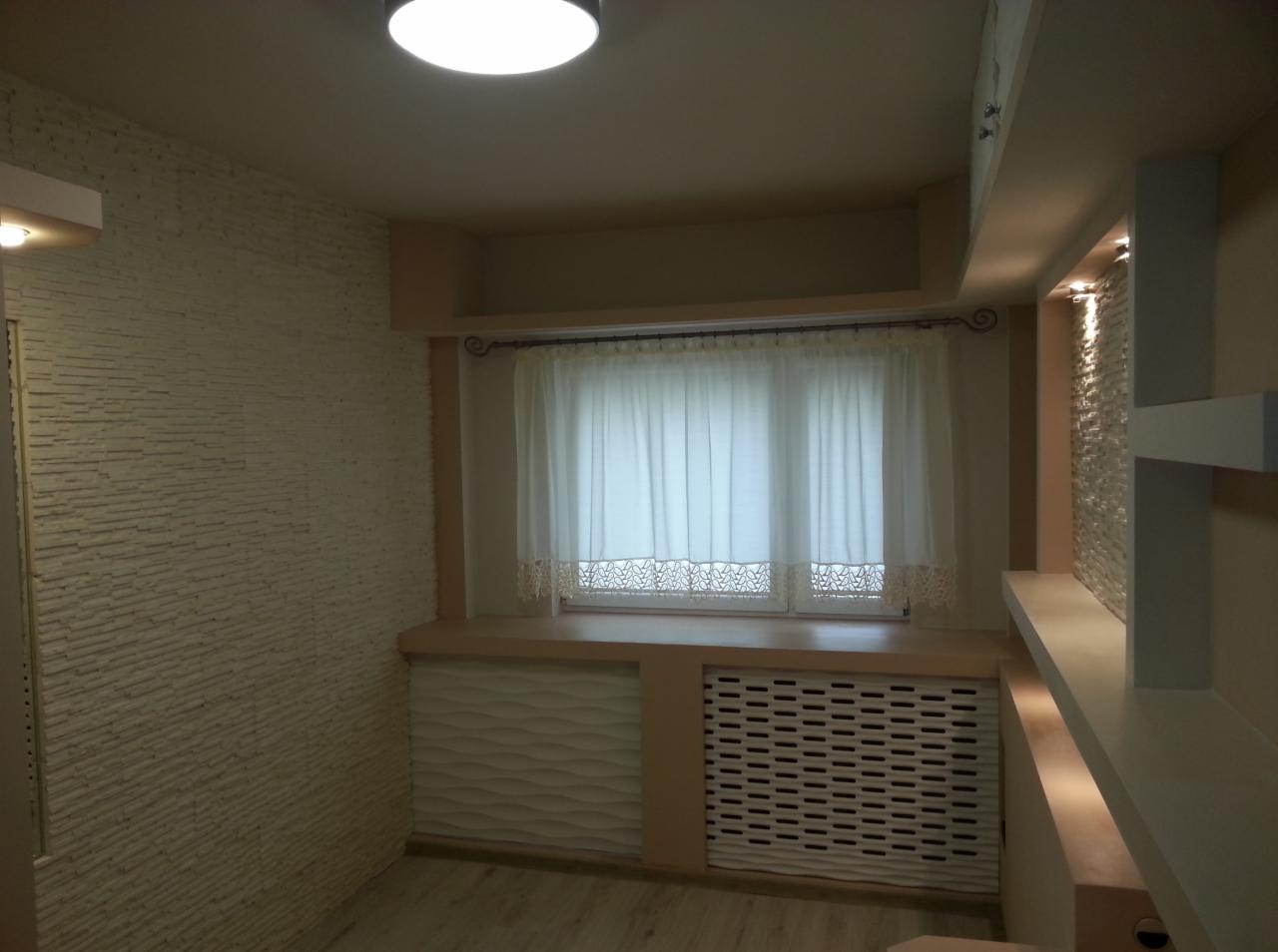 Jasne wnętrze pokoju po remoncie, widok na okno z firanką, dekoracyjne panele ścienne z efektem 3D oraz zabudowa grzejnika z ażurowym frontem.