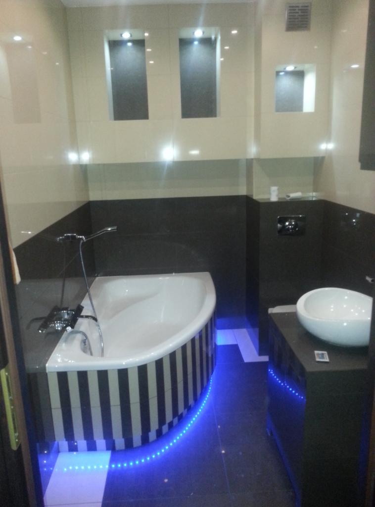 Nowoczesna łazienka z wanną w czarno-białe pasy podświetloną niebieskim LED, umywalką na ciemnej szafce oraz wnękami w ścianie z oświetleniem punktowym.