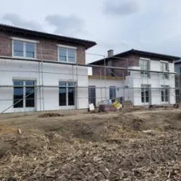 Domy jednorodzinne w trakcie prac elewacyjnych, widoczne rusztowania i ocieplenie styropianem na frontowej ścianie pierwszego budynku, częściowo wykończona elewacja z cegły klinkierowej na piętrze,...