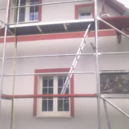 Ściana domu z białym tynkiem i czerwoną ramą wokół okien, widoczna spod rusztowania i drabiny, z satelitą na dachu.