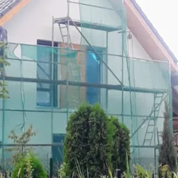 Dom jednorodzinny w trakcie prac elewacyjnych, osłonięty siatką na rusztowaniu, widoczny fragment dachu z ciemnej dachówki, bujna roślinność w ogródku przed domem.