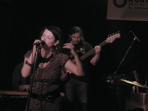 Wokalistka Asia Kwaśnik śpiewa do mikrofonu podczas koncertu z zespołem; basista w tle, klawiszowiec po lewej, scena oświetlona punktowo.