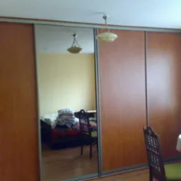 Brązowa szafa przesuwna z lustrem, odbicie pokoju z łóżkiem i krzesłem, widok z boku na stół z koronkowym obrusem.