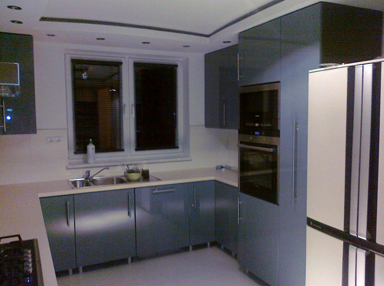 Nowoczesna kuchnia w odcieniach szarości z błyszczącymi frontami szafek, widoczne okno z żaluzjami, zlewozmywak i piekarnik w zabudowie.