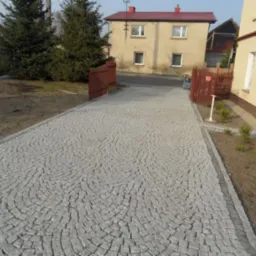 Podjazd z jasnoszarej kostki granitowej ułożonej w półkoliste wzory, obrzeża z ciemniejszej kostki, w tle dom jednorodzinny z czerwonym dachem i brązowym ogrodzeniem.