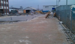 Układanie kostki brukowej we Wrocławiu: widok na plac budowy z częściowo ułożoną kostką, przygotowane podłoże i stosy kostki w tle, w oddali budynek i maszyny budowlane.