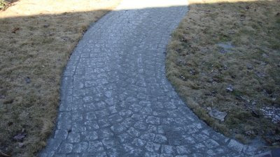 Zakrzywiony chodnik z szarej kostki granitowej, otoczony suchą trawą, widziany z góry.