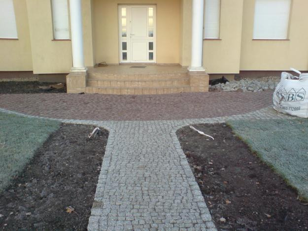 Ułożona ścieżka z szarej kostki granitowej rozwidlająca się przed wejściem do domu z beżową elewacją, w tle schody z brązowej kostki, po prawej stronie widoczny worek z materiałami budowlanymi.
