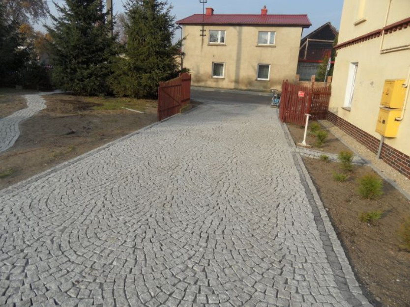 Podjazd z jasnoszarej kostki granitowej ułożonej w półkoliste wzory, obrzeża z ciemniejszej kostki, w tle dom jednorodzinny z czerwonym dachem i brązowym ogrodzeniem.