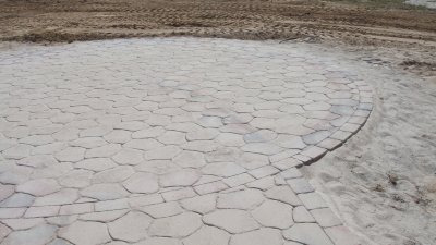 Okrągły plac wyłożony nieregularnymi, jasnymi kamieniami, obrzeże z prostokątnych kostek, widoczne ślady opon na ziemi w tle.