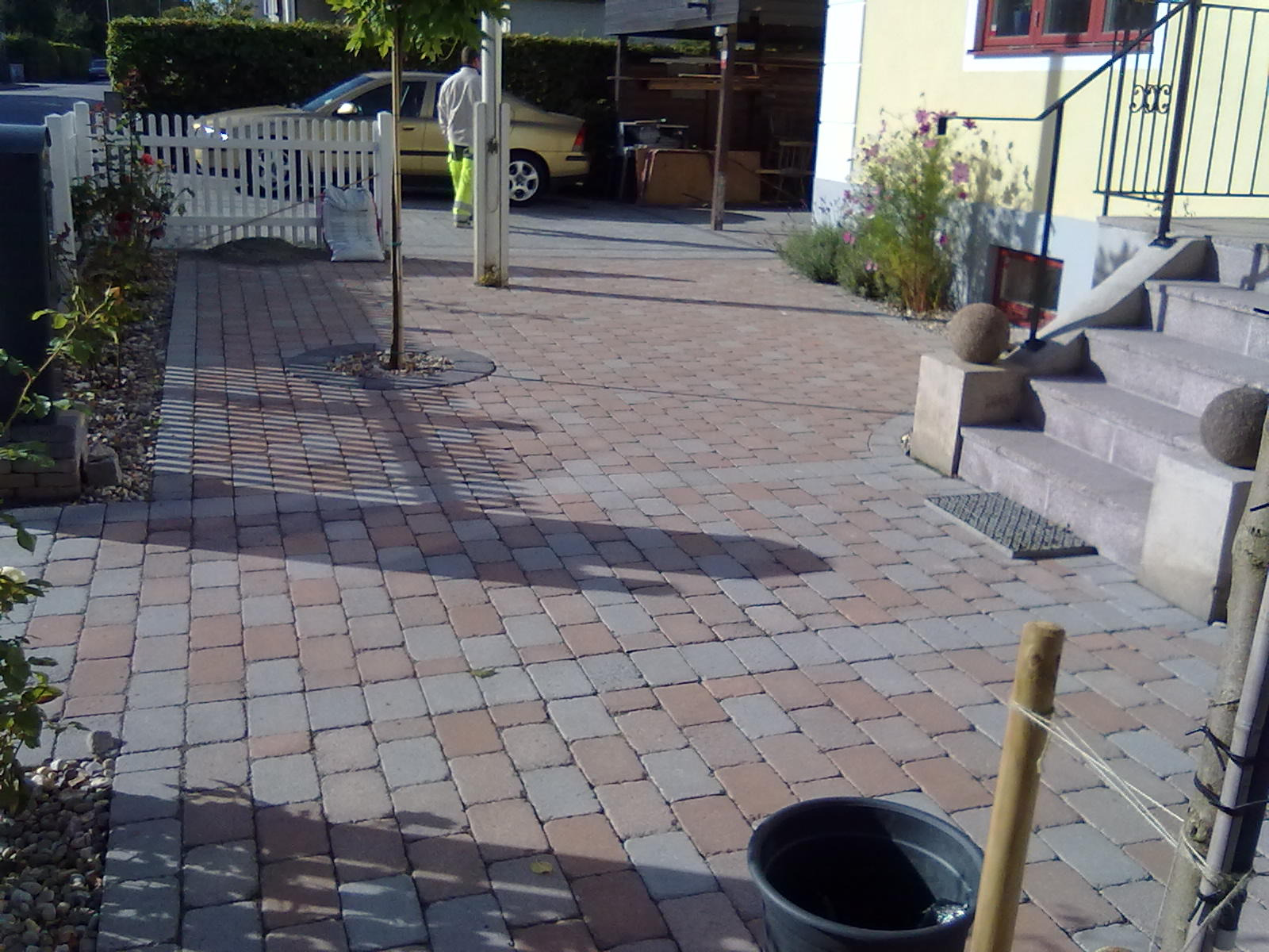 Ułożona kostka brukowa w odcieniach szarości i czerwieni tworząca ścieżkę prowadzącą do domu z jasną elewacją i schodami, widoczne drzewko w okrągłej oprawie z kamieni, w tle biały płot...