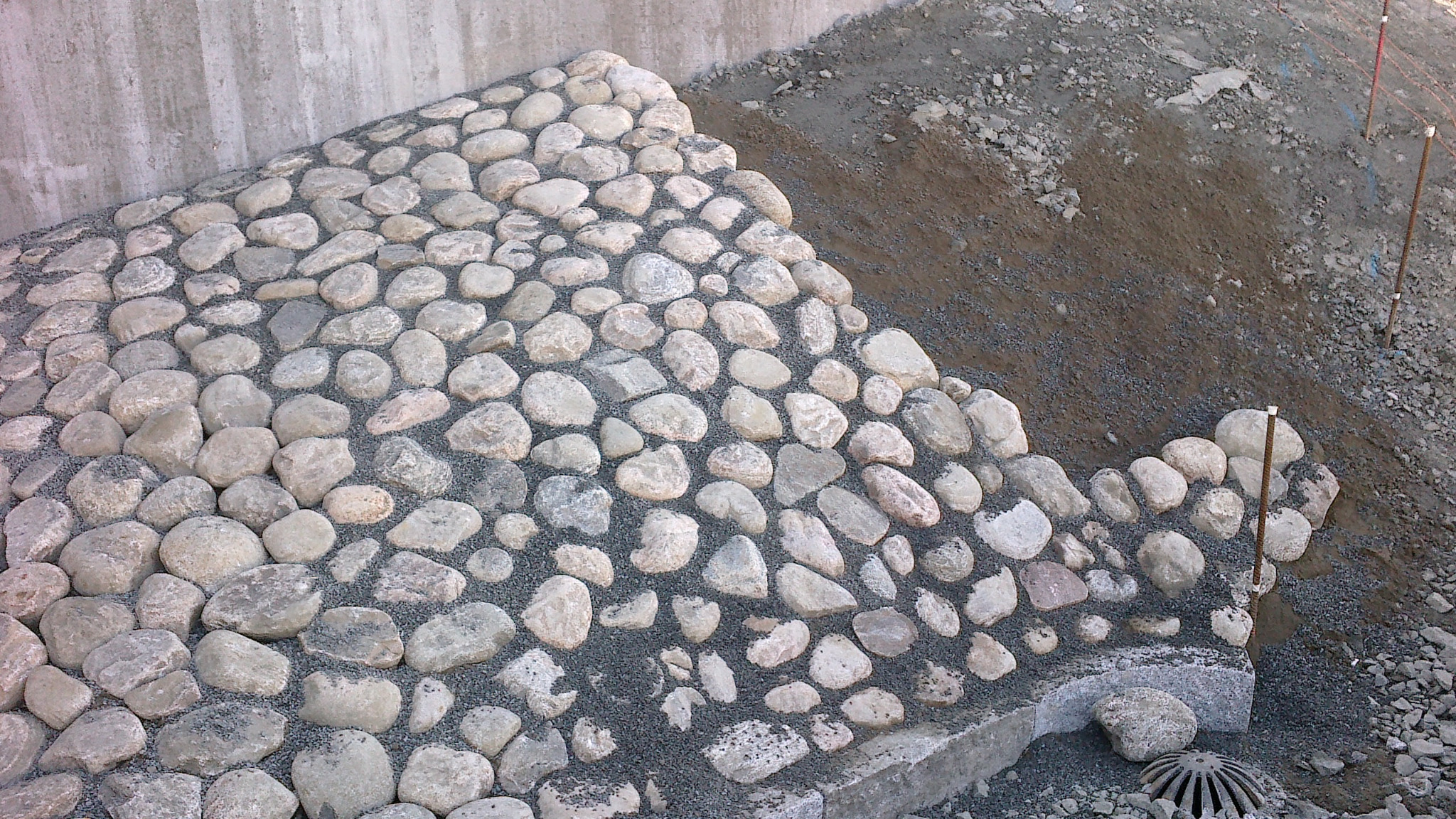 Skarpa umocniona okrągłymi kamieniami z szarą podsypką, widoczne także elementy instalacji odwadniającej i ziemia z gruzem w tle.