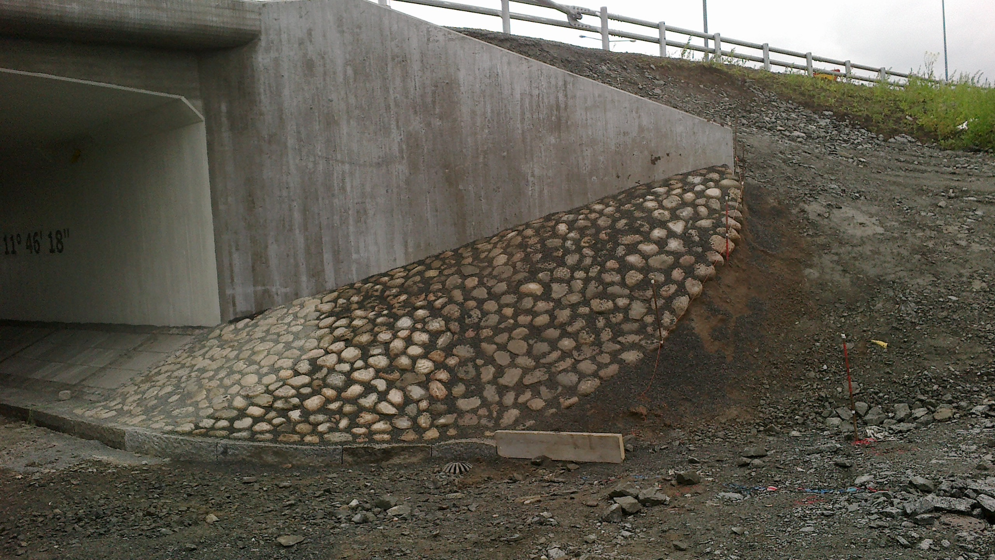 Skarpa zabezpieczona okrągłymi kamieniami przy betonowym tunelu drogowym, widoczne geodezyjne znaczniki w gruncie.