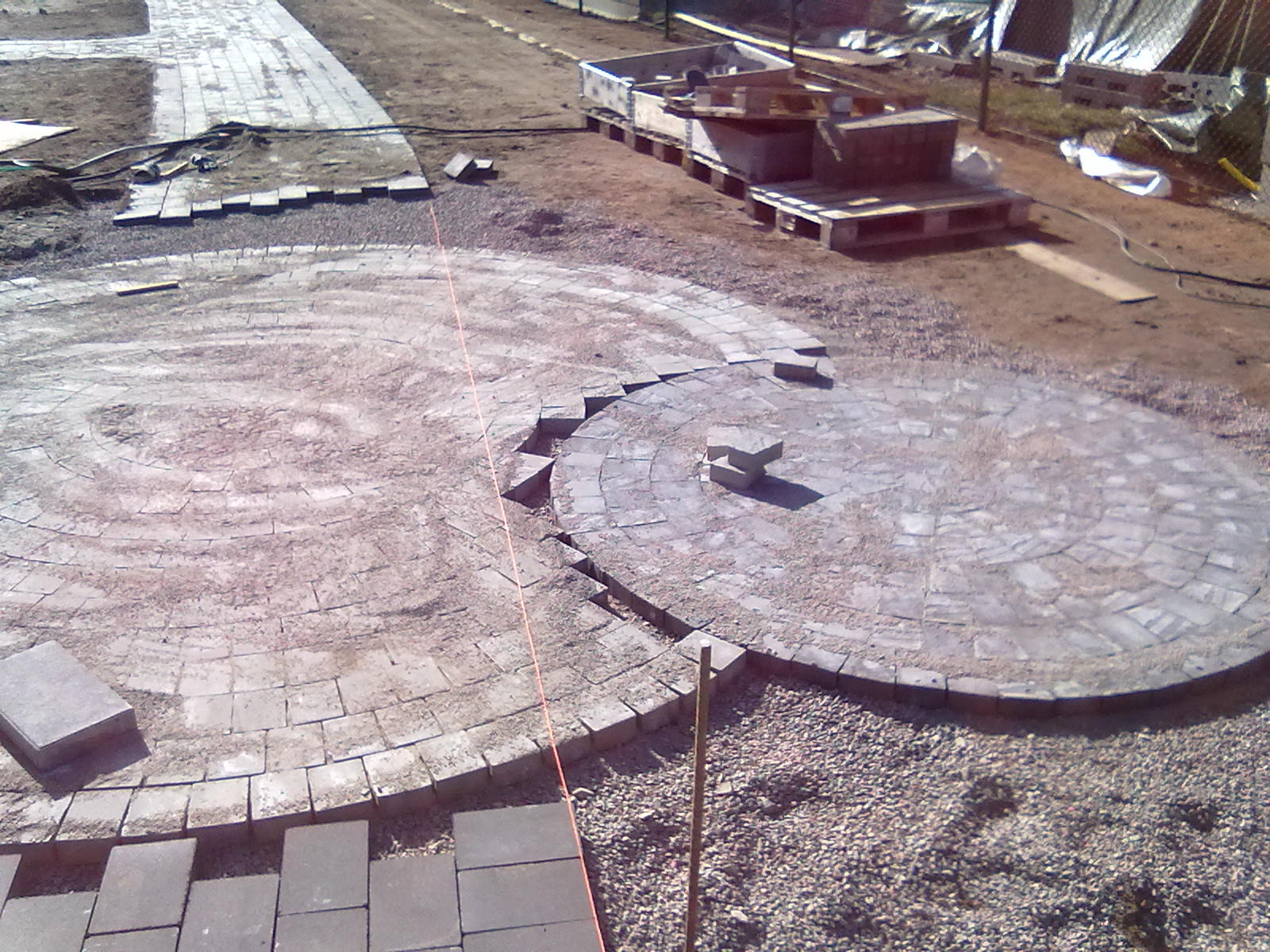 Widok z góry na plac budowy z częściowo ułożoną kostką brukową w kształcie koła i ścieżki, widoczne palety z materiałami budowlanymi w tle.